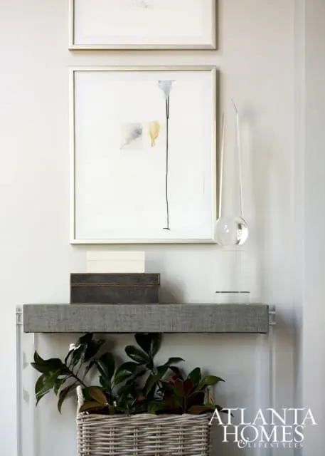 Gray console table with stacked boxes, abstract art, and a clear glass sculpture. A woven basket with green plants sits below.