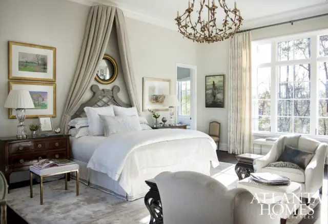 Serene bedroom with a large white bed draped in a taupe canopy, flanked by a dark wood nightstand and a plush armchair. A large window overlooks a wooded landscape.