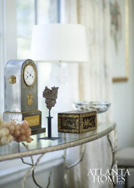 Silver antique clock with gold accents, a bust on a stand, and a decorative box on a glass-top console table with a lamp and grapes