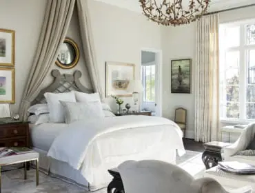 Bedroom with a draped canopy bed, white bedding, ornate chandelier, and large windows overlooking trees.