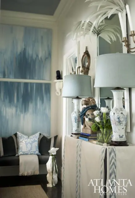Hallway with abstract blue and white art, dark upholstered bench with patterned pillows, and a console table displaying blue and white ginger jars, lamps, and a floral arrangement.