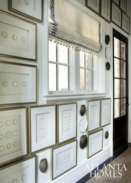 Hallway with gallery wall of framed art featuring circular motifs, decorative shells, and a roman shade window with pom-pom trim.