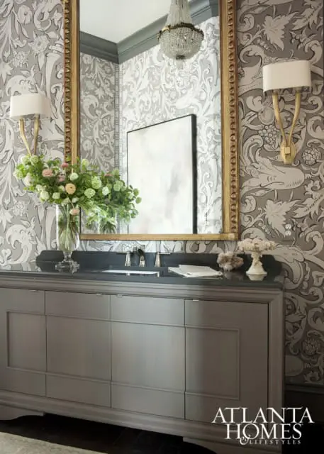 Gray vanity with dark countertop and ornate gold mirror above. Walls feature gray and white damask wallpaper. A crystal chandelier hangs overhead.