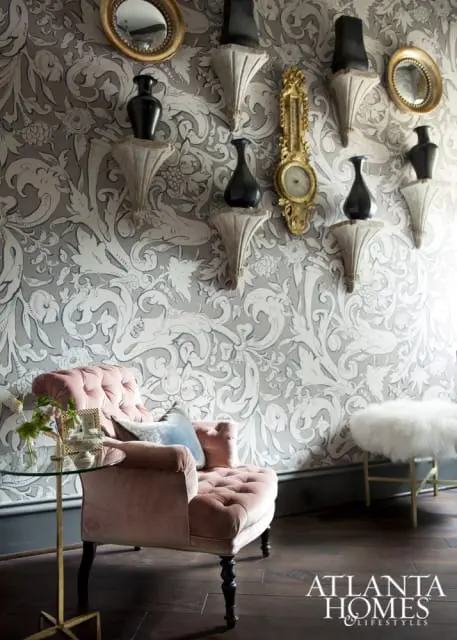 Pink velvet tufted armchair next to a glass side table with a vase of flowers. The wall features a grey and white damask wallpaper with decorative shelves holding black vases and a gold clock.