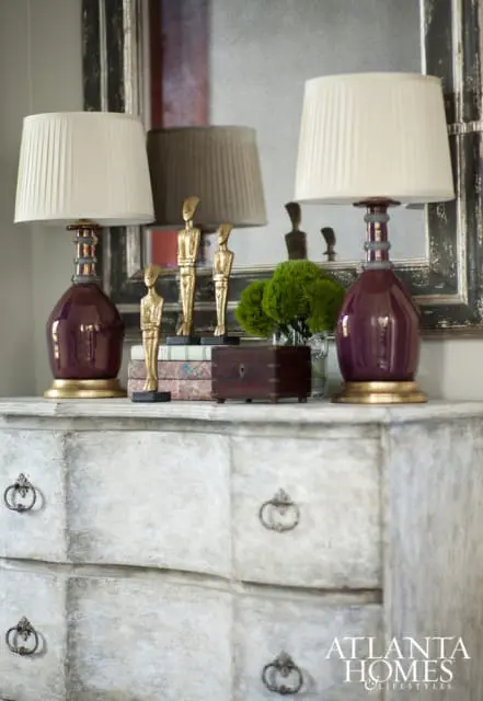 Two purple glass lamps with gold bases and cream pleated shades flank gold figurines on a distressed white dresser, reflected in a large mirror.