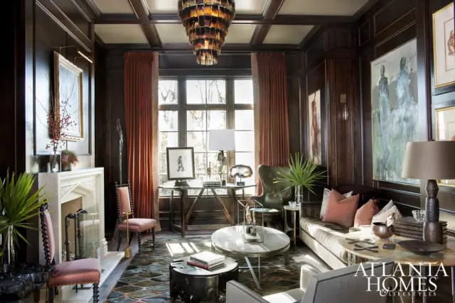 Dark wood paneled library with a white marble fireplace, terracotta drapes, leather armchair, and a sectional sofa.