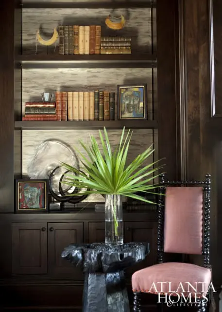 Dark wood built-in bookshelves filled with antique books, artwork, and decorative objects. A glass vase with palm fronds sits on a carved wood side table next to a chair with a salmon-colored seat and back.