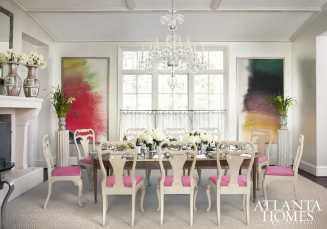 Formal dining room with a large wooden table set for a meal, surrounded by ornate cream-colored chairs with pink upholstered seats. Abstract art adorns the walls, and a crystal chandelier hangs overhead.