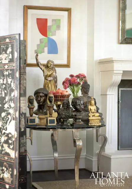 Round glass-top table displaying an assortment of busts, statues, and a bowl of pomegranates, with a framed abstract artwork and a decorative screen in the background.