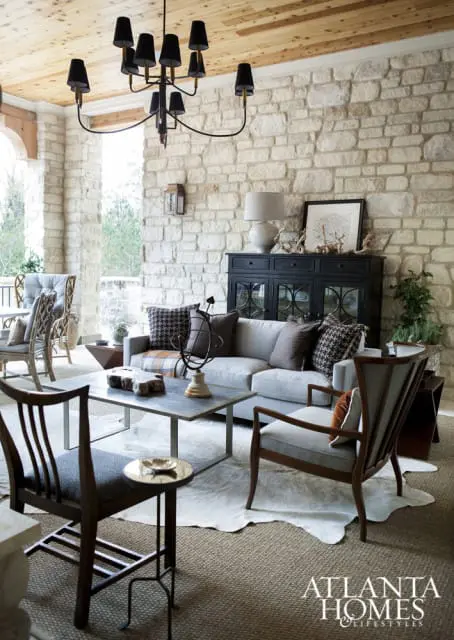 Covered porch living area with stone walls, wood plank ceiling, neutral sofa and chairs, metal coffee table, and a black chandelier.
