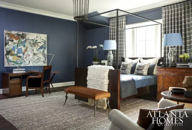 Bedroom with navy blue walls, a dark wood four-poster bed with blue bedding, a leather bench, a wooden desk with a chair, and abstract art.