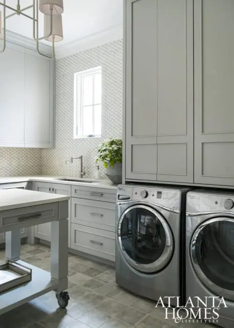 Gray laundry room with stacked washers and dryers, white geometric tile backsplash, and a rolling island with a stone countertop.