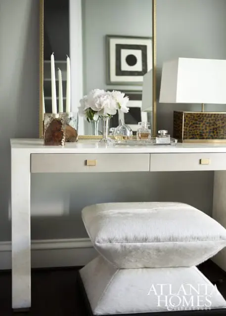 White vanity table with gold hardware, topped with a mirror, candles, peonies, perfume bottles, and a leopard print lamp. A white fur stool sits in front.
