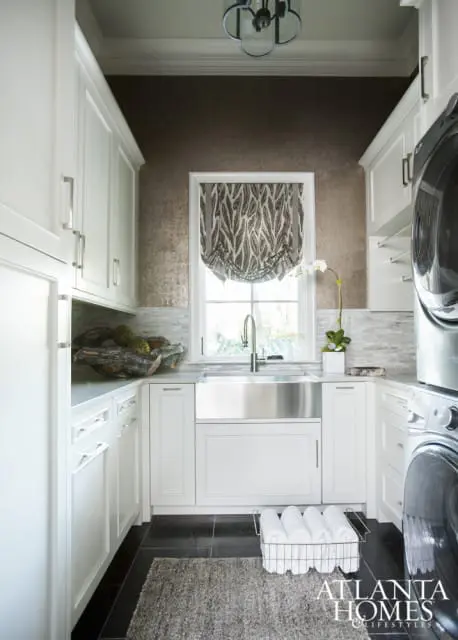 White laundry room with white shaker cabinets, stainless steel farmhouse sink, dark tile flooring, and a metallic textured wallpaper above the backsplash.