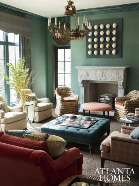 Green living room with tufted teal ottoman, red sofa, upholstered chairs, ornate chandelier, and fireplace with carved mantel.