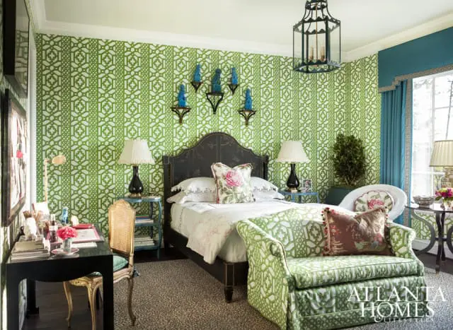 Bedroom with green and white geometric wallpaper, a dark wood bed with floral pillows, a black desk with a cane-backed chair, and a green patterned sofa.