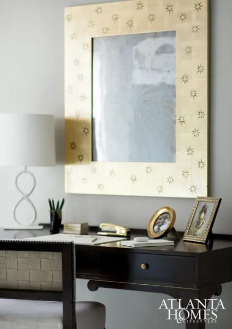 Dark wood desk with gold starburst mirror above, white lamp, framed photos, and stapler.
