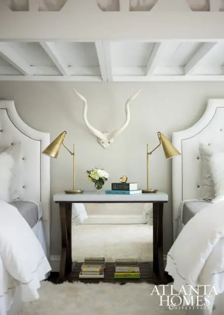 Bedroom with two white tufted headboards, marble-topped nightstand with brass lamps, and white faux fur rug. A white animal skull with horns hangs on the wall.
