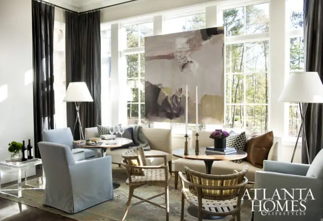 Living room with light blue slipcovered armchairs, woven accent chairs, neutral upholstered seating, and a large abstract painting above a round table with candles and flowers.