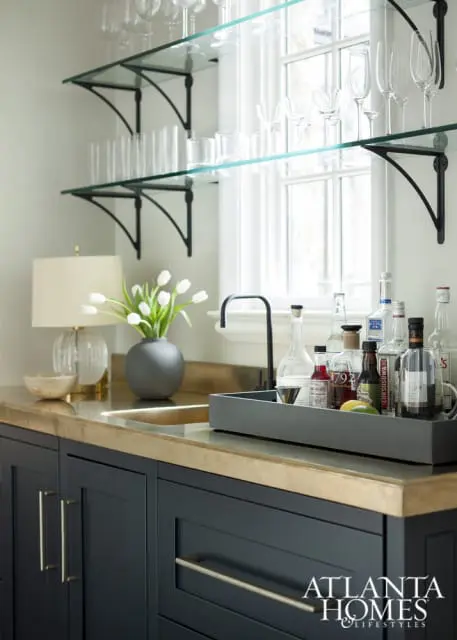Dark blue bar cabinets with brass hardware topped with a brass countertop. A gray tray holds liquor bottles and garnishes next to a black faucet. Glass shelves above display various glassware.