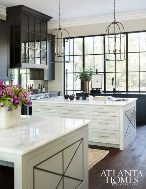 Luxury kitchen with white marble island, dark gray cabinetry, and large grid windows overlooking trees. Two cage pendant lights hang above the island.