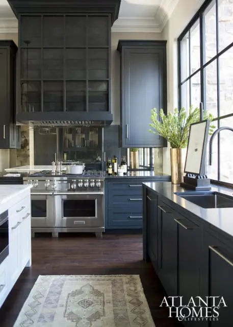 Dark blue kitchen with stainless steel range, mirrored backsplash, and large black-framed window. A patterned rug lies on the dark wood floor.