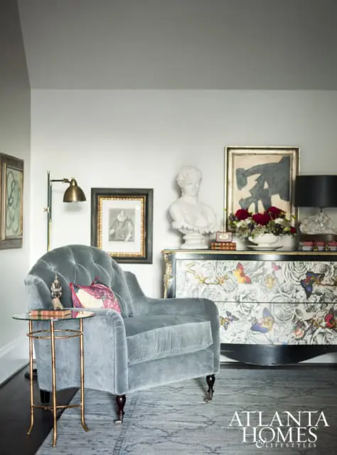 Gray tufted armchair next to a side table with a glass top and brass frame. Behind the chair is a dresser with a butterfly and floral pattern, topped with a bust, artwork, and flowers.