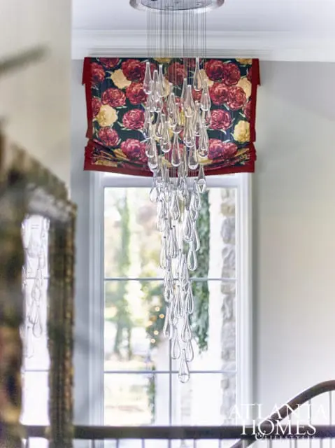Clear teardrop chandelier hanging in front of a window with floral Roman shades and a stone accent wall.