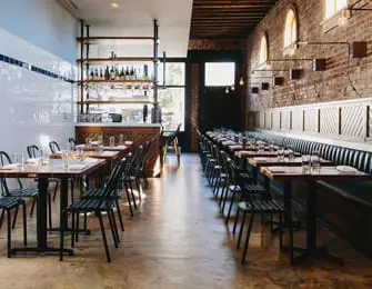 Restaurant dining room with exposed brick walls, long banquette seating, and tables set with place settings.