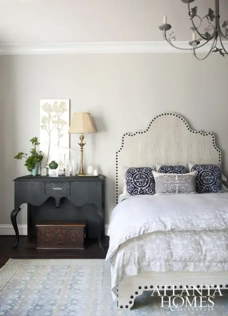 Bedroom with a light wood headboard adorned with nailhead trim, white bedding, and decorative pillows. A dark wood console table with a lamp and artwork sits beside the bed.