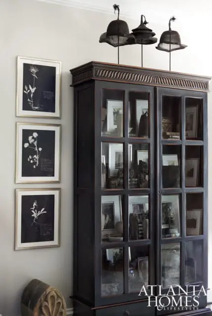Dark wood curio cabinet with glass doors displaying framed botanical prints and decorative objects, next to a wall with three framed botanical prints.