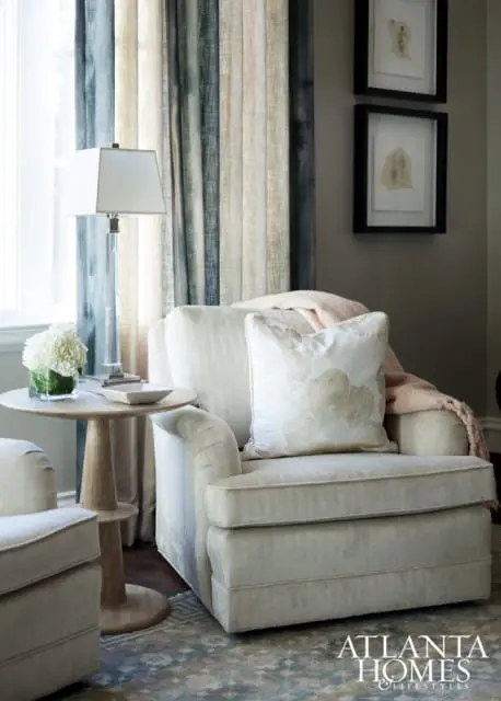 Cream upholstered armchair with a floral accent pillow and pink throw blanket, next to a round wooden side table with a glass vase of white flowers and a small dish.