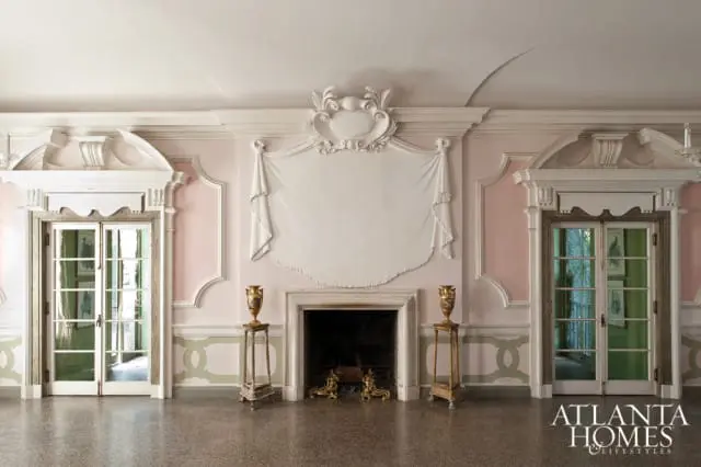 Elegant living room with pink walls, ornate white molding, a fireplace with decorative urns, and double doors with glass panes.