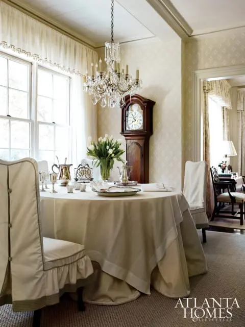 Dining room with a round table covered in a beige tablecloth, a grandfather clock, and a crystal chandelier.