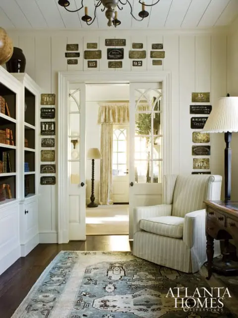 Living room with white paneled walls, built-in white bookshelves, a striped armchair, a patterned rug, and antique signs displayed on the walls.