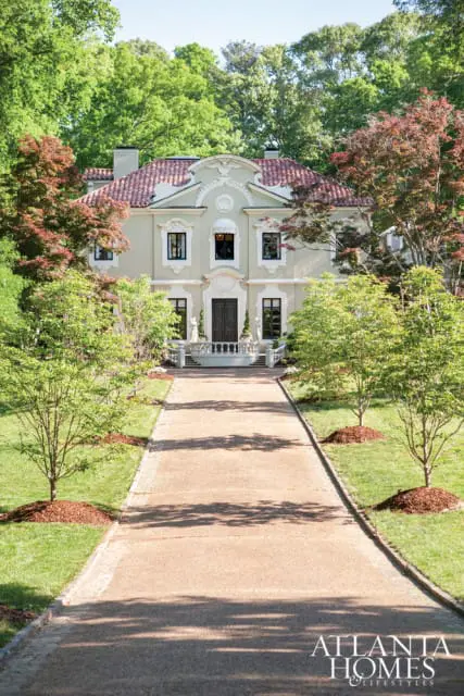 Light green stucco house with red tile roof, ornate facade, and double front doors, approached by a gravel driveway lined with trees and manicured lawns.