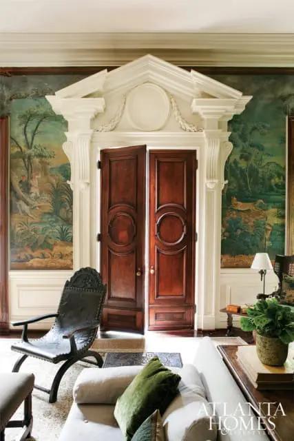 Grand entryway with double mahogany doors, ornate white pediment, and scenic wallpaper depicting a forest landscape. A dark leather chair and plush sofa are visible in the foreground.