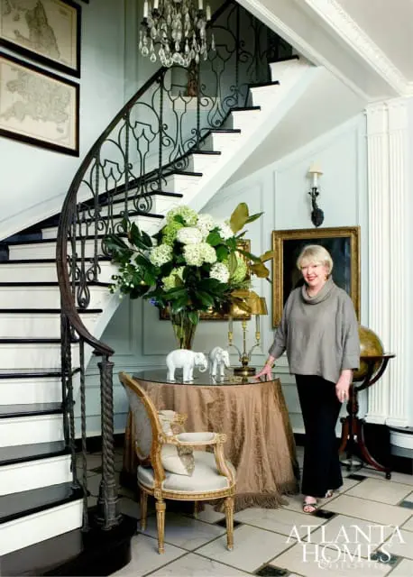 Woman standing next to a round table with a floral arrangement and elephant figurines in a foyer with a curved staircase and chandelier.