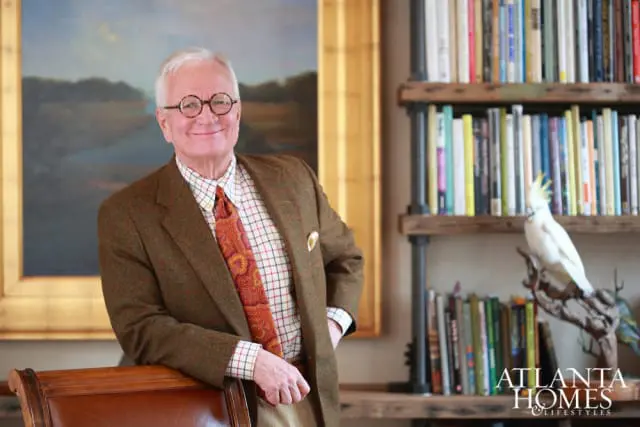 Man with glasses wearing a brown tweed jacket, plaid shirt, and patterned tie, leaning on a wooden chair. Behind him is a painting and a bookshelf with a white cockatoo figurine.