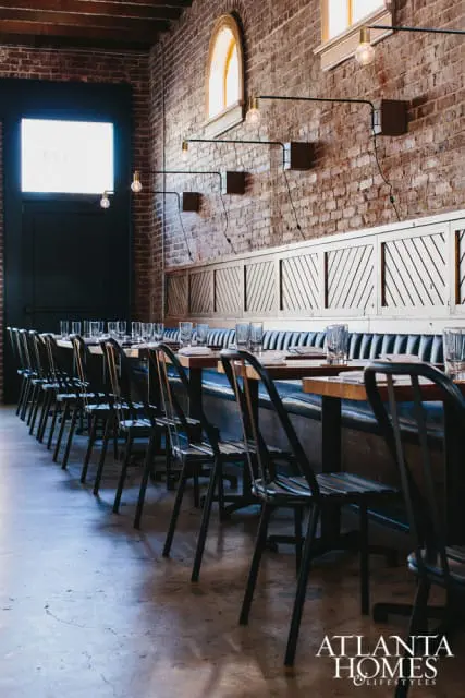 Restaurant dining room with long banquette seating, dark metal chairs, and exposed brick walls. Industrial pendant lights hang above wooden tables set with glassware.