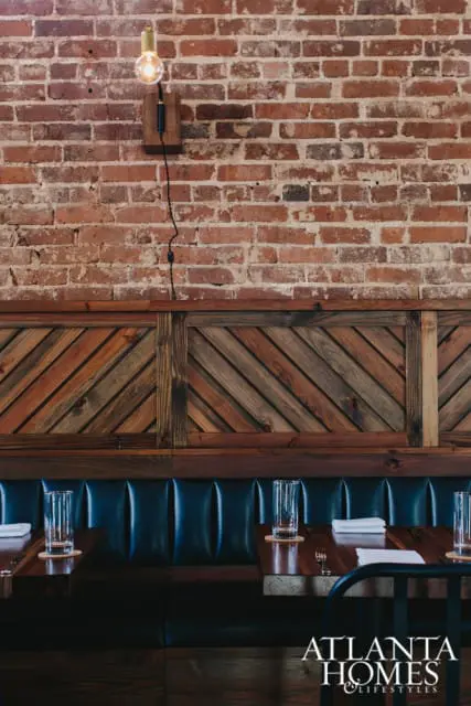 Restaurant interior with exposed brick wall, dark wood paneling, and navy blue tufted booths. A single Edison bulb light fixture hangs on the wall.