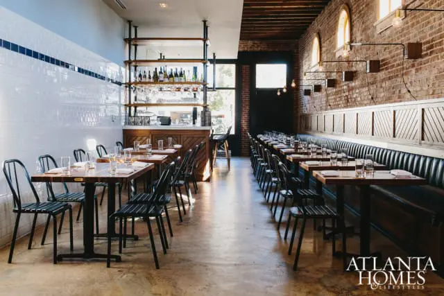 Restaurant interior with white subway tile walls, wooden tables, black metal chairs, and a brick accent wall with banquette seating.