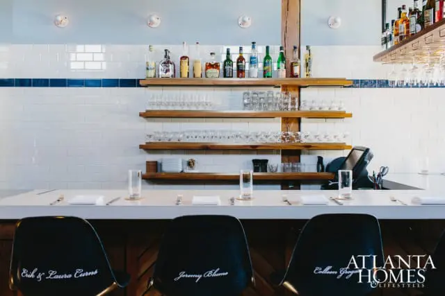 Bar area with white subway tile backsplash, wooden shelves stocked with bottles and glassware, and a white countertop with place settings. Black chairs with names inscribed are in the foreground.
