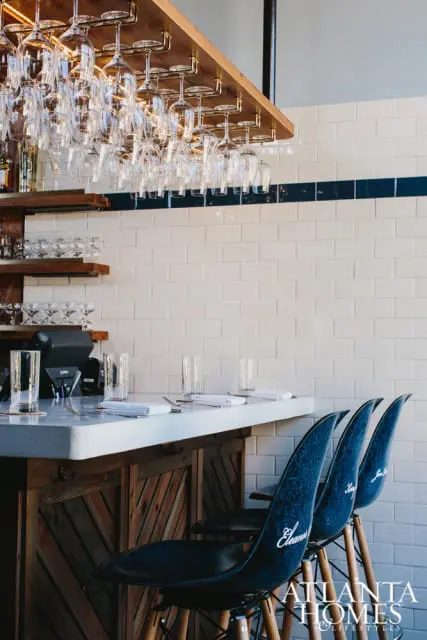 Bar area with white subway tile backsplash, concrete countertop, and dark blue bar stools with white lettering. A wooden rack with hanging wine glasses is above the bar.