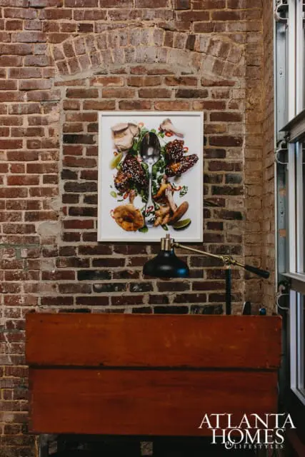 Framed artwork of food, including chicken wings, mushrooms, and ginger, hangs on a brick wall above a wooden desk with a black desk lamp.
