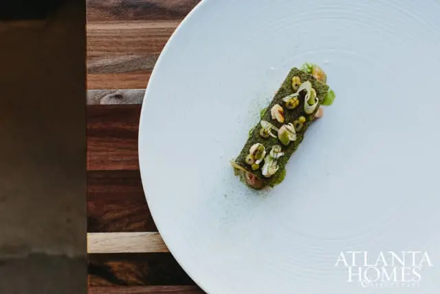Gourmet dish with green topping and small white garnishes on a white plate, next to a wooden surface.