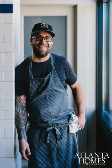 Man wearing a baseball cap, glasses, and a striped apron, with a tattooed arm leaning against a tiled wall.