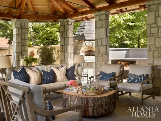 Covered outdoor living space with stone pillars, wooden ceiling, sectional sofa and chairs with blue and striped pillows, and a rustic log coffee table.