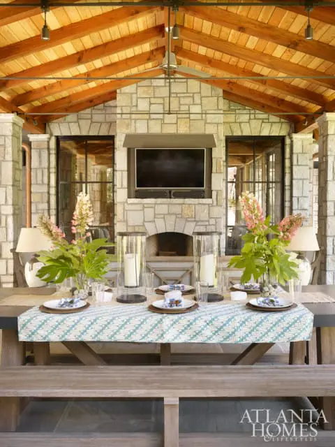 Covered outdoor dining area with stone fireplace, mounted television, and a table set for a meal with floral arrangements.