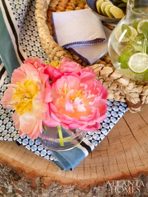 Pink peonies in a glass vase on a wooden surface with a pitcher of lime water and patterned napkins in the background.
